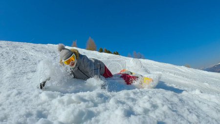 Foto de Female snowboarder falls after losing balance while making a snowboard turn. Young woman lands into snow after losing balance at snowboarding down the slope at snowy mountain ski resort on sunny day. - Imagen libre de derechos