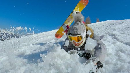 Téléchargez les photos : Young woman sliding through soft snow after she falls at snowboarding. Female beginner snowboarder is sliding face forward after falling down on a snowy ski slope at mountain ski resort. - en image libre de droit