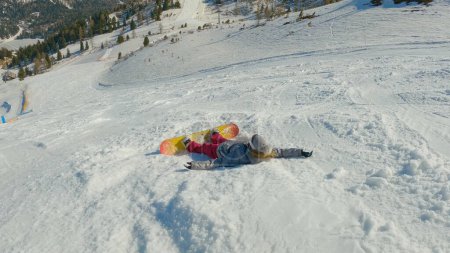 Foto de Girl snowboarder falls back on snow while learning to snowboard on ski slope. Young woman landing in snow after losing balance while snowboarding down the piste at snow-covered mountain ski resort. - Imagen libre de derechos