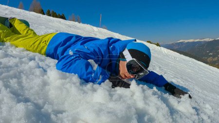 Foto de Man crashes into snow while learning to snowboard on snowy ski slope. Young male snowboarder tries to learn how to turn and ride snowboard at mountain ski resort on a beautiful sunny day. - Imagen libre de derechos