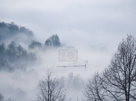 Foto de Tall construction crane rising from the winter mist rolling over countryside. Metal crane among treetops and foggy clouds. Modern building machinery above construction site hidden in the dense fog. - Imagen libre de derechos