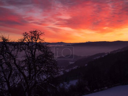 Téléchargez les photos : Majestic play of glowing sunset colors above snow-covered hilly countryside. Stunning vista of vibrant atmospheric mood above rural landscape. Serene view from the hilltop over snowy valley at sundown - en image libre de droit