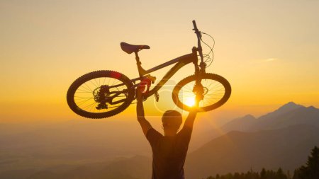 Téléchargez les photos : CLOSE UP, LENS FLARE : Un homme excité lève son vélo au-dessus de sa tête après une amusante balade à vélo de fond. Dans toute la Slovénie. Un homme méconnaissable soulève son vélo dans les airs au coucher du soleil doré pittoresque. - en image libre de droit