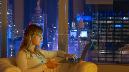 Photo for CLOSE UP DOF: Bright lights of Times Square flash below the Caucasian girl working on her computer late at night. Young businesswoman does research on computer while sitting on the couch in her room. - Royalty Free Image