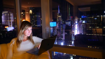 Foto de CLOSE UP, COPY SPACE: Mujer trabajando en su portátil desde su habitación de hotel en lo alto de Times Square. Joven mujer de negocios caucásica trabajando horas extras desde su oficina con una impresionante vista del centro de Nueva York. - Imagen libre de derechos