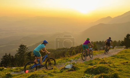 Téléchargez les photos : LENS FLARE : Quatre jeunes voyageurs descendent leur VTT au lever du soleil. Touristes en vélo électrique sur un sentier étroit dans les montagnes verdoyantes slovènes idylliques lors d'une soirée d'été ensoleillée. - en image libre de droit