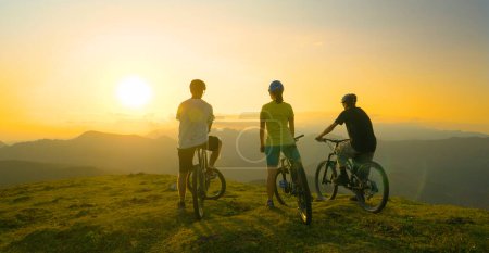 Photo for SUN FLARE: Three unrecognizable friends observe the evening nature from a mountain top after a fun mountain bike journey in Slovenia. Mountain cyclists watch the sunrise before a fun downhill race. - Royalty Free Image