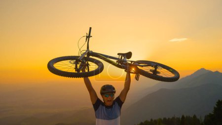 Téléchargez les photos : FERMER, SOLEIL FLARE : Heureux homme caucasien crie tout en tenant son vélo au-dessus de sa tête au coucher du soleil. Touriste masculin joyeux célèbre gagner une course de vélo de fond lors d'une soirée d'été ensoleillée. - en image libre de droit