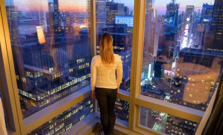 Téléchargez les photos : FERMER : Jeune femme regardant les rues et les gratte-ciel de la ville métropolitaine aux États-Unis par une matinée ensoleillée. Touriste méconnaissable se tient dans la chambre d'hôtel et observe Times Square à l'aube. - en image libre de droit