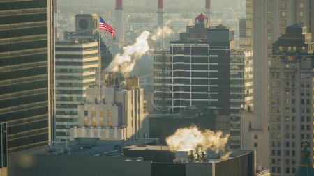 Foto de CERRAR: En la soleada mañana en Nueva York se emite al aire un espeso humo blanco procedente de chimeneas industriales y rascacielos. Edificios de oficinas modernos de gran altura y naturaleza contaminante de fábrica con humos dañinos - Imagen libre de derechos
