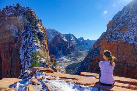 Téléchargez les photos : LENS FLARE : La voyageuse s'assoit et observe le canyon enneigé de Angel's Landing. Jeune femme s'assoit après une randonnée et regarde le magnifique canyon de couleur rouge dans le parc national de Zion dans l'Utah. - en image libre de droit