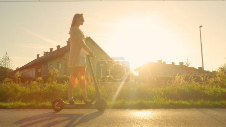 Foto de LOW ANGLE, LENS FLARE: Mujer joven monta un scooter eléctrico más allá de la ciudad suburbana al atardecer. Millennial chica en tacones altos disfruta de un divertido paseo a través de los suburbios en un scooter de alta tecnología al amanecer de oro - Imagen libre de derechos