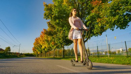 Foto de ÁNGULO BAJO: Mujer caucásica feliz en tacones negros divirtiéndose montando una scooter eléctrica por la avenida escénica en los suburbios. Sonriendo millennial paseos e-scooter por carretera corriendo más allá de los árboles de color otoño - Imagen libre de derechos