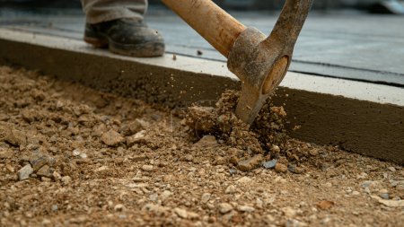 Photo for CLOSE UP, DOF: Unrecognizable worker uses a pickaxe to start digging into the soft gravel ground next to a building under construction. Pickaxe strikes into soil mixed with gravel. Construction site - Royalty Free Image