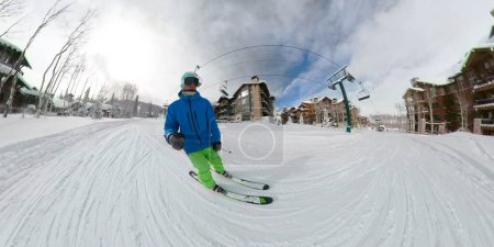Foto de DESCRIPCIÓN SELFIE Turista masculino disfruta esquiando a lo largo de la pista arreglada y bajo el telesilla pasando por los hoteles de la estación de esquí. Cruceros en esquiador por la suave pendiente de una pista para principiantes en Copper Mountain - Imagen libre de derechos