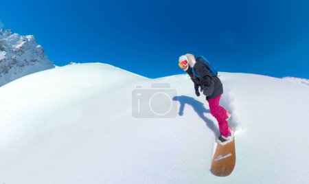 Téléchargez les photos : SELFIE : Une jeune femme sourit alors qu'elle fait de l'héliport dans les montagnes Rocheuses ensoleillées de la Colombie-Britannique. Joyeux snowboarder féminin pulvérise la poudre de champagne fraîche tout en chevauchant hors piste par une journée ensoleillée. - en image libre de droit