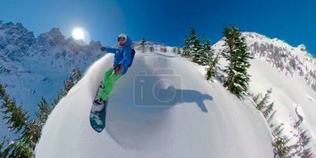 Foto de SELFIE, LENS FLARE: El joven heliboarder rocía la nieve fresca mientras se esculpe por la montaña en la soleada Columbia Británica. Impresionante toma de acción de una trituradora freeride snowboarder en las Montañas Rocosas. - Imagen libre de derechos