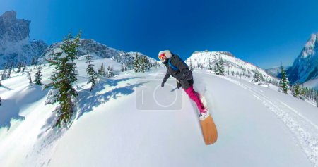 Téléchargez les photos : SELFIE : Joyeux snowboarder féminin sculpte hors piste dans les pittoresques Rocheuses canadiennes. Femme caucasienne joyeuse profite d'une journée d'hiver ensoleillée en faisant un voyage d'héliport cool en Colombie-Britannique. - en image libre de droit