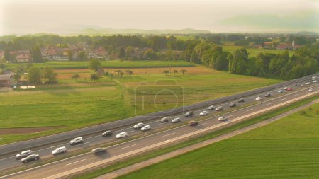 Photo for DRONE: Golden afternoon sunbeams shine on vehicles stuck in a jam on the freeway crossing the idyllic countryside. Commuters and tourists move through a congestion on the highway caused by road works. - Royalty Free Image