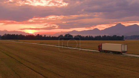 Photo for AERIAL, COPY SPACE: Flying along a freight truck hauling a white cargo container across the picturesque countryside at sunrise. Lorry hauls merchandise across the rural landscape on a sunny evening. - Royalty Free Image