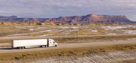 AERIAL: Velocidades de camiones semirremolque a lo largo de la autopista que cruza el desierto en Utah. Paisaje invernal del desierto rodea camión que conduce por la carretera interestatal que atraviesa escarpado desierto de EE.UU..