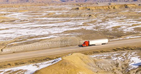 AERIAL: Barren wintry landscape surrounds a cargo truck driving across wintry Utah. Red semi-trailer truck hauls a heavy container across the snowy desert. Truck driving down the interstate freeway.