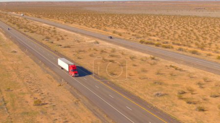 AERIAL: Red cargo truck hauls a heavy container across the barren landscape. Aerial shot of a semi-trailer truck transporting merchandise across the rugged Utah desert on a sunny spring afternoon.