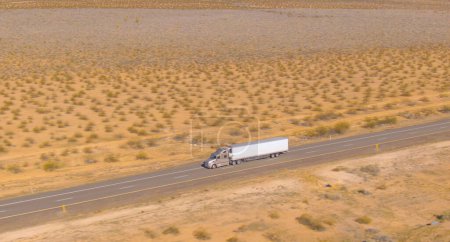 Téléchargez les photos : AÉRIAL : Prise de vue cinématographique d'un camion cargo alors qu'il descend la pittoresque autoroute asphaltée traversant le désert accidenté. Le camion transporte un lourd conteneur dans la nature sauvage. Semi-remorque camion traversant le désert - en image libre de droit