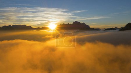 Foto de DRONE, SOL FLARE: Volando sobre las nubes y una cresta rocosa en lo alto de los Alpes franceses al amanecer. Los rayos dorados del sol de la tarde iluminan la cordillera y las nubes se reúnen sobre el campo francés. - Imagen libre de derechos