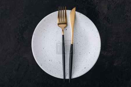 Photo for Empty plate with fork and knife on dark stone background. Gold and black tableware with white plate, top view, copy space - Royalty Free Image