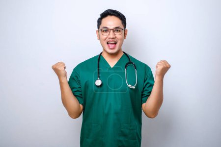 Photo for Excited asian male doctor wearing scrubs and eyeglasse doing winning gesture congrats or praise someone isolated over white background - Royalty Free Image