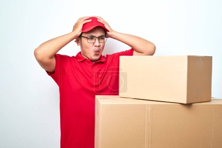 Téléchargez les photos : Courrier asiatique inquiet et troublé en uniforme rouge et tête de capture de chapeau et haletant choqué regardant les boîtes en carton isolées sur fond blanc - en image libre de droit