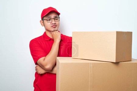 Téléchargez les photos : Courrier asiatique inquiet et troublé en uniforme rouge et tête de capture de chapeau et haletant choqué regardant les boîtes en carton isolées sur fond blanc - en image libre de droit