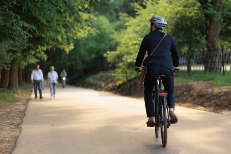 Photo for Stylish young man in black suit cycling on bike on paved road in summer. Back view of unrecognizable businessman in protective helmet riding on bicycle through park area. Concept of eco lifestyle. - Royalty Free Image