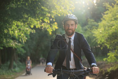 Téléchargez les photos : Happy attractive businessman in helmet riding by bicycle on way to work in morning. Front view of confident man driving bike, while looking at camera against sunlight. Concept of eco lifestyle. - en image libre de droit