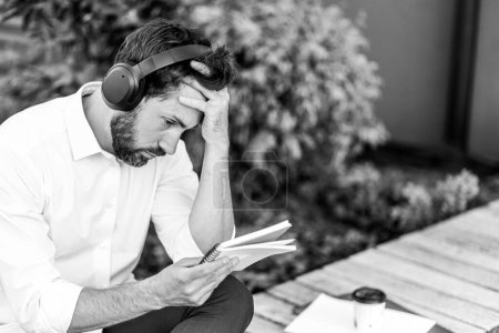 Foto de Thoughtful man in headphones checking notes, while sitting on bench outdoors. Monochrome portrait of pensive guy with hand on head, thinking, looking at notebook in park, copyspace. Concept of work. - Imagen libre de derechos