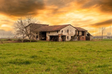 Altes verlassenes italienisches Bauernhaus mit typischer ländlicher Architektur der Poebene in der Provinz Cuneo, Italien, bei Sonnenuntergang mit farbigem bewölkten Himmel