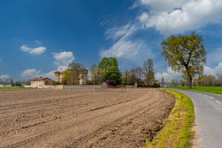 Po Valley countryside landscape with road leading to Galateri di Suniglia Castle, Cuneo, Italy among plowed fields and blue sky with white cloud