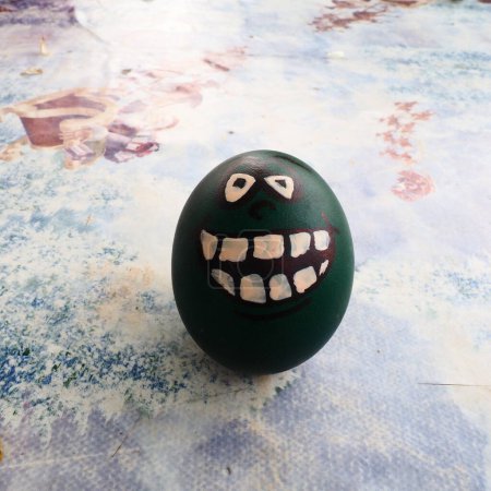 Photo for Easter egg dyed green with a painted laughing face. Cool grimace with eyes and big white teeth. Scary mug for Halloween. Emoticon for Easter. Light abstract background - Royalty Free Image