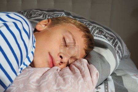 Foto de Un hermoso chico caucásico de 10 años de edad con el pelo rubio, vestido con pijamas a rayas, duerme en una cama con una manta esponjosa, abrazando una almohada. Luz suave del sol de la mañana fluye a través de la ventana - Imagen libre de derechos
