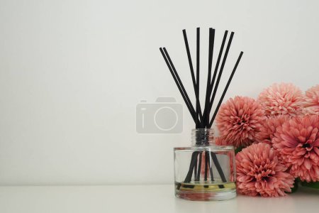 Composition with incense sticks, essential oil in a glass vase and pink terry artificial flowers in the interior of a white room. White background. Lots of flowers and scent sticks. Copy space.