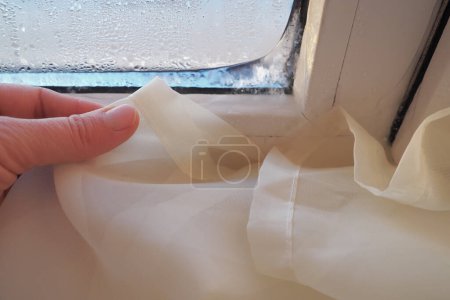 Photo for Curtain - white tulle froze to pvc window frame. Melting ice on the glass pane. Problems of heating, maintaining an ergonomic climate in the apartment. Mold on windows. Womans hand. Economic crisis. - Royalty Free Image
