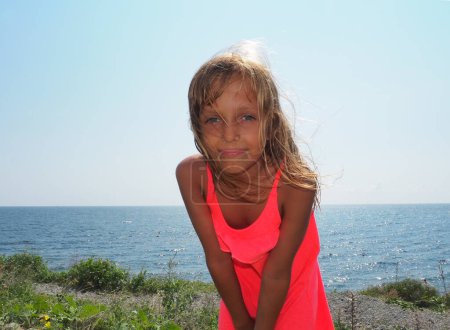 Foto de Una jovencita guapa de 8 años de edad con el pelo largo y rubio, la piel bronceada sonríe en el contexto de un paisaje marino. Vestido de vestido rosa. Los rayos de sol iluminan el cabello de los niños. Cielo azul y horizonte - Imagen libre de derechos