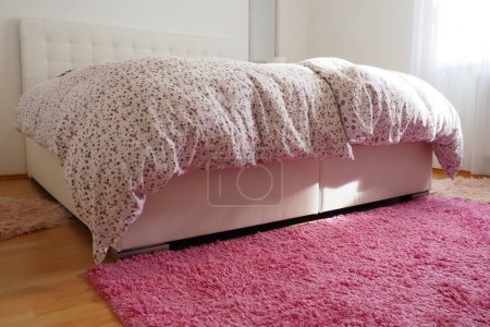 Téléchargez les photos : Chambre compacte confortable avec des meubles blancs tapis rose et rideaux de tulle. Couverture de plumes moelleuse est jeté sur le lit. Bonjour romantique. Lit double avec tête de lit rembourrée. Lumière de la fenêtre - en image libre de droit