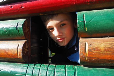A 10-year-old Caucasian boy with blond hair and gray eyes looks out of the window of a wooden house on a childrens playground. Peekaboo. The child hid. Pretty face.