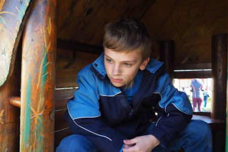 Un niño caucásico de 10 años con el pelo rubio y los ojos grises mira por la puerta de una casa de madera en un parque infantil. Peekaboo. El niño se escondió. Bonita cara. El chico está sentado en una casita.