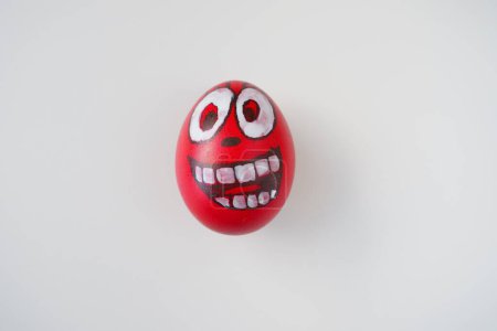 Photo for Easter egg painted red, white and black with a cheerful toothy face. Boiled chicken dyed egg with red shell. White background. Big eyes, white teeth on the fun face. Copy space. - Royalty Free Image