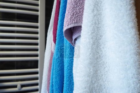 Towels hang next to a heated towel rail, wall radiator or radiator. White, blue, pink, red towels. Organization of household items in the bathroom. Housekeeping