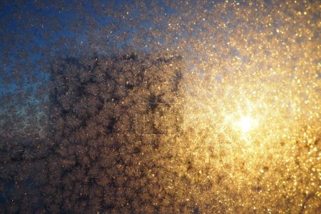 Das frostige Muster auf Fensterglas entsteht durch die Kondensation von Wasserdampf auf Glas, das unter 0 Grad abgekühlt ist. Frostblumen, Locken und Kristalle. Morgen gelb-orange tief stehende Wintersonne am Horizont.