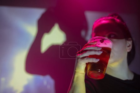 Téléchargez les photos : Femme tenant et buvant une tasse de bière en plastique dans la boîte de nuit. - en image libre de droit